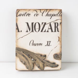 Könnte beinhalten: Rechteckige dekorative Plakette mit dem Text "Maitre de Chapelle A. MOZART Oeuvre XI." Die Plakette hat ein gealtertes Aussehen mit einem Goldblatt-Design am unteren Rand. Der Hintergrund ist cremefarben.