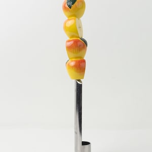 Può includere: Un levatorsoli per mele con manico in acciaio inossidabile e parte superiore decorativa con quattro mele stilizzate. Le mele sono dipinte di giallo e rosso con accenti di foglie verdi, conferendo un design stravagante a tema frutta.