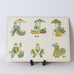 May include: A rectangular placemat with a cream background featuring six illustrated scenes of people at a market. The drawings are rendered in shades of green and yellow, depicting figures with produce, fish, and umbrellas, all framed by a thin green border.