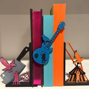 May include: A set of decorative bookends featuring a cleaver, a blue guitar, and a circus tent. The bookends are in front of colorful books. The guitar has a skull and silhouette design. The cleaver has a pink bird and spiderweb design.