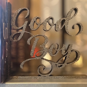 May include: A decorative metal bookend in a textured, dark gray finish. The bookend features the words "Good Boy" in an elegant script. The design is suitable for holding books on a shelf.
