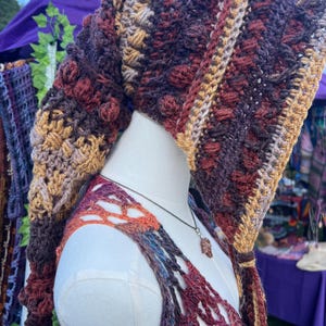 May include: A long, pointed crochet hood featuring a textured bobble stitch pattern in variegated shades of deep burgundy, dark brown, and golden tan. The hood drapes down the back and includes long, dangling ties at the front edges with matching yarn.