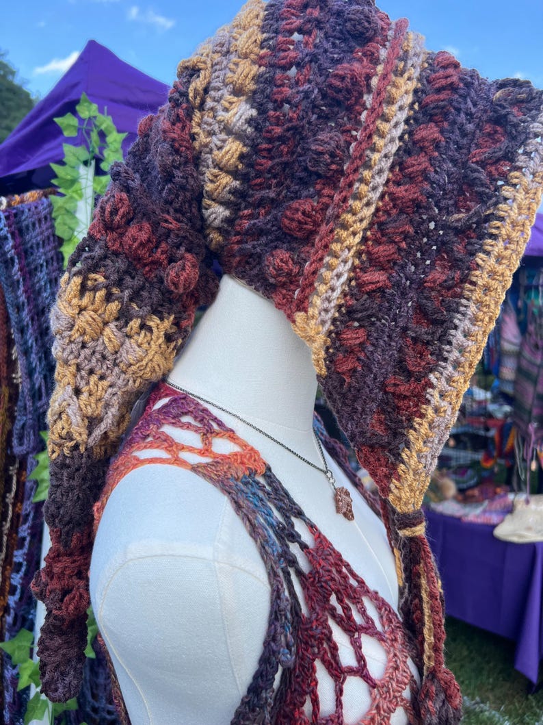 May include: A long, pointed crochet hood featuring a textured bobble stitch pattern in variegated shades of deep burgundy, dark brown, and golden tan. The hood drapes down the back and includes long, dangling ties at the front edges with matching yarn.