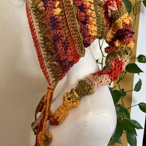 May include: A long, pointed crochet hood featuring a variegated color palette of burnt orange, olive green, deep purple, and cream. The textured knit displays alternating patterns of bobble stitches and vertical stripes, ending in long, dangling braided ties.