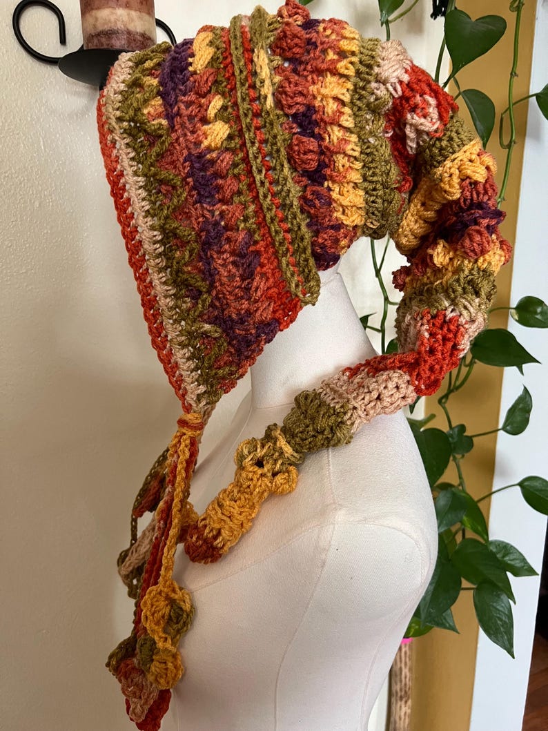 May include: A long, pointed crochet hood featuring a variegated color palette of burnt orange, olive green, deep purple, and cream. The textured knit displays alternating patterns of bobble stitches and vertical stripes, ending in long, dangling braided ties.
