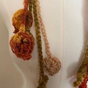 May include: A crochet garland featuring multiple long, thin, braided strands in shades of olive green, burnt orange, mustard yellow, and cream. Each strand terminates in a small, circular, textured crochet flower or leaf-like motif.