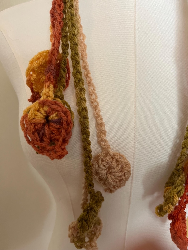 May include: A crochet garland featuring multiple long, thin, braided strands in shades of olive green, burnt orange, mustard yellow, and cream. Each strand terminates in a small, circular, textured crochet flower or leaf-like motif.