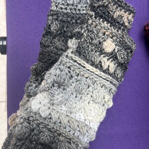 May include: A pair of handmade crochet fingerless gloves in shades of gray, white, and black. The gloves feature a textured stitch pattern and a rolled cuff. The yarn appears to be a soft, wool blend. The gloves are displayed on a purple surface.