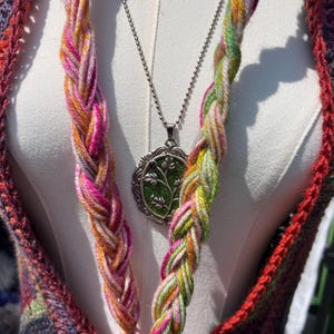 May include: A silver-toned pendant necklace featuring an oval frame with a green textured inlay and a small purple accent, worn with two thick, braided yarn strands in variegated pink, yellow, and green hues over a dark multicolored crochet garment.