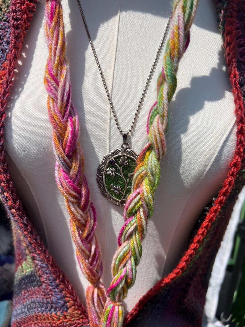 May include: A silver-toned pendant necklace featuring an oval frame with a green textured inlay and a small purple accent, worn with two thick, braided yarn strands in variegated pink, yellow, and green hues over a dark multicolored crochet garment.