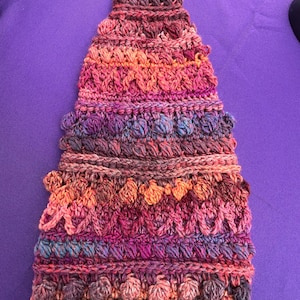 May include: A handmade, colorful crochet hat in a cone shape. The hat features a variety of textured stitches and a gradient of colors, including shades of orange, pink, purple, and brown. The hat is displayed on a purple surface.
