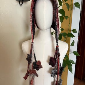 May include: A hooded head covering crafted from variegated yarn in shades of brown, rust, gray, and cream. The hood features long, dangling braided ties on each side, each adorned with multiple small, crocheted leaf-shaped embellishments.