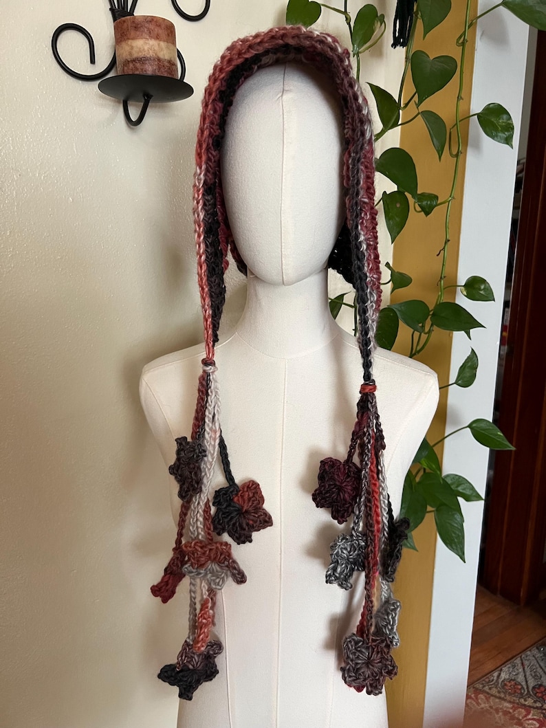 May include: A hooded head covering crafted from variegated yarn in shades of brown, rust, gray, and cream. The hood features long, dangling braided ties on each side, each adorned with multiple small, crocheted leaf-shaped embellishments.