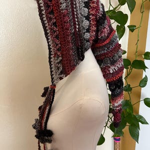 May include: A long, pointed crochet hood featuring a textured pattern of bobble stitches and stripes in shades of burgundy, charcoal gray, light gray, and white. The hood includes long, dangling braided ties finished with small, decorative crochet flower motifs.