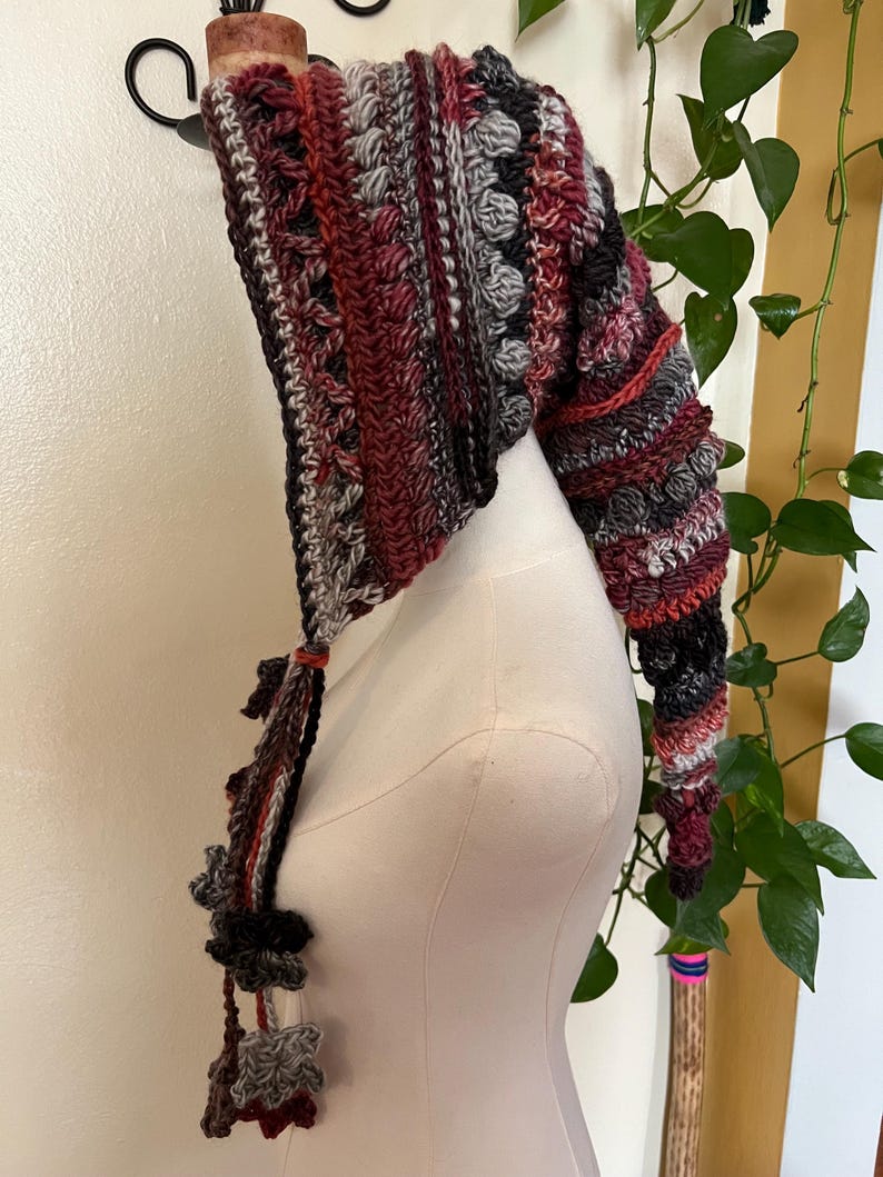 May include: A long, pointed crochet hood featuring a textured pattern of bobble stitches and stripes in shades of burgundy, charcoal gray, light gray, and white. The hood includes long, dangling braided ties finished with small, decorative crochet flower motifs.