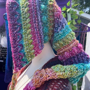 May include: A multi-colored crochet hood featuring a vibrant blend of purple, teal, green, and pink yarn. The hood displays a textured pattern with bobble stitches and a long, braided tassel hanging from the front edge, worn on a mannequin.