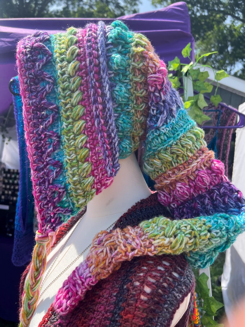May include: A multi-colored crochet hood featuring a vibrant blend of purple, teal, green, and pink yarn. The hood displays a textured pattern with bobble stitches and a long, braided tassel hanging from the front edge, worn on a mannequin.