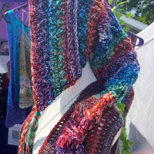 May include: A hooded vest crafted from multicolored yarn featuring a vibrant blend of blue, purple, red, and green tones. The garment displays a textured crochet pattern with raised bobble stitches and long, braided ties hanging from the hood.