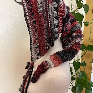 May include: A long, pointed crochet hood featuring a variegated color palette of deep burgundy, dark charcoal, muted grey, and burnt orange. The textured design incorporates alternating rows of bobble stitches and ribbed patterns with long, dangling braided ties.