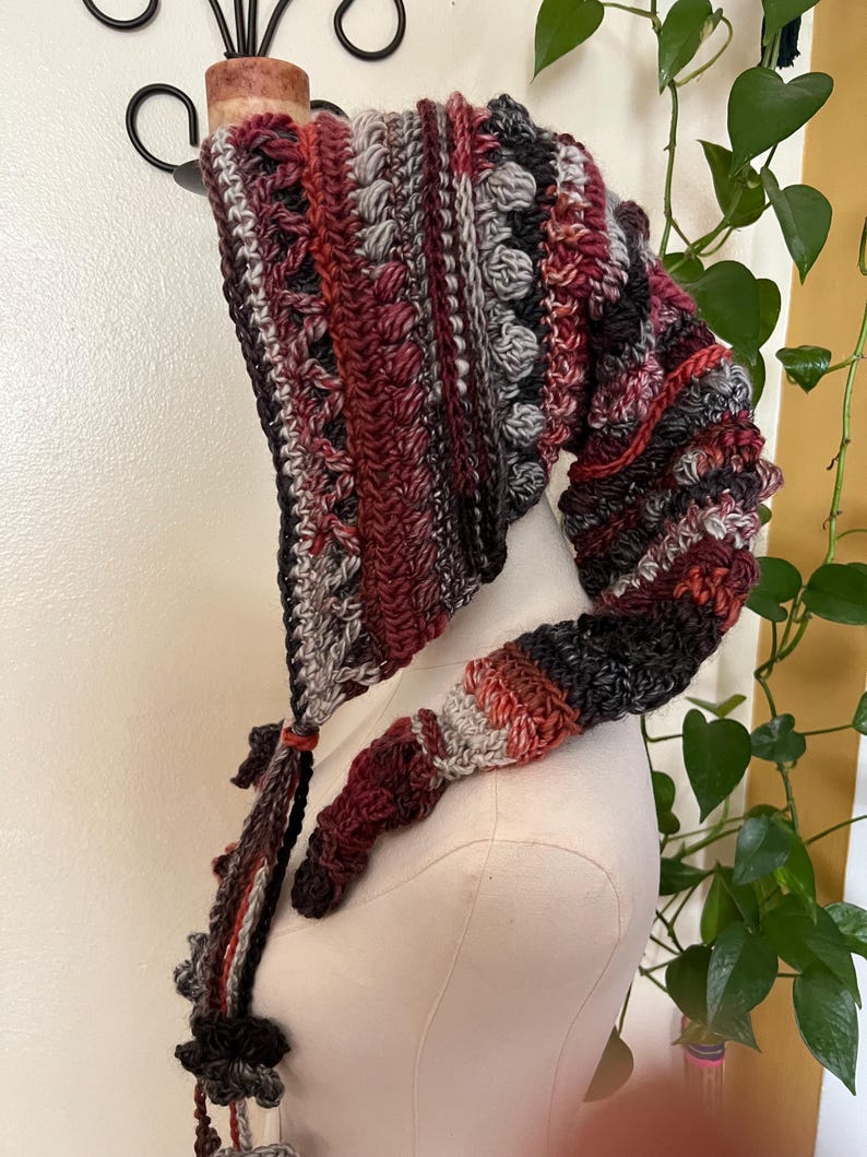 May include: A long, pointed crochet hood featuring a variegated color palette of deep burgundy, dark charcoal, muted grey, and burnt orange. The textured design incorporates alternating rows of bobble stitches and ribbed patterns with long, dangling braided ties.