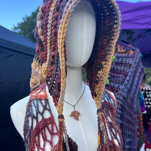 May include: A hooded crochet garment featuring a variegated color palette of deep burgundy, burnt orange, mustard yellow, and brown. The piece has an open, net-like stitch pattern and is displayed on a mannequin wearing a small copper-colored leaf pendant.