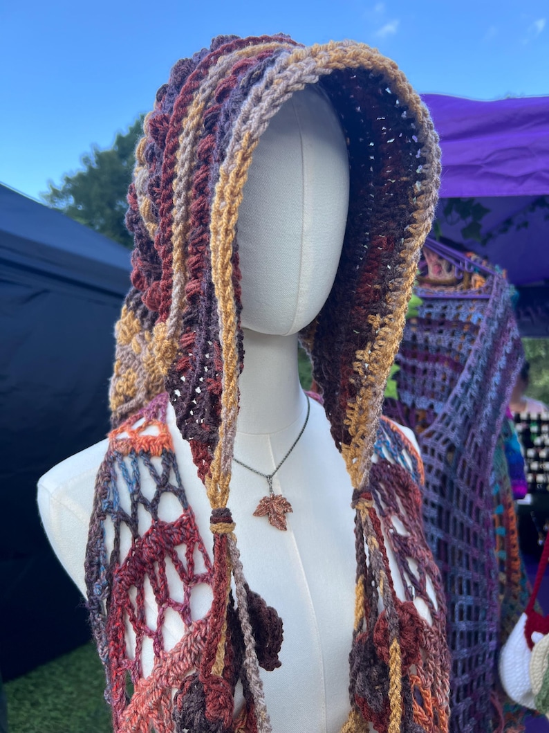 May include: A hooded crochet garment featuring a variegated color palette of deep burgundy, burnt orange, mustard yellow, and brown. The piece has an open, net-like stitch pattern and is displayed on a mannequin wearing a small copper-colored leaf pendant.