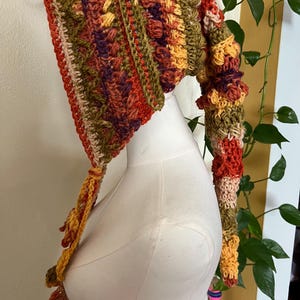 May include: A long, pointed crochet hood featuring a textured, multi-colored pattern in shades of orange, olive green, deep purple, and cream. The headpiece has a tapered, elongated tail and includes braided ties hanging from the front edges.