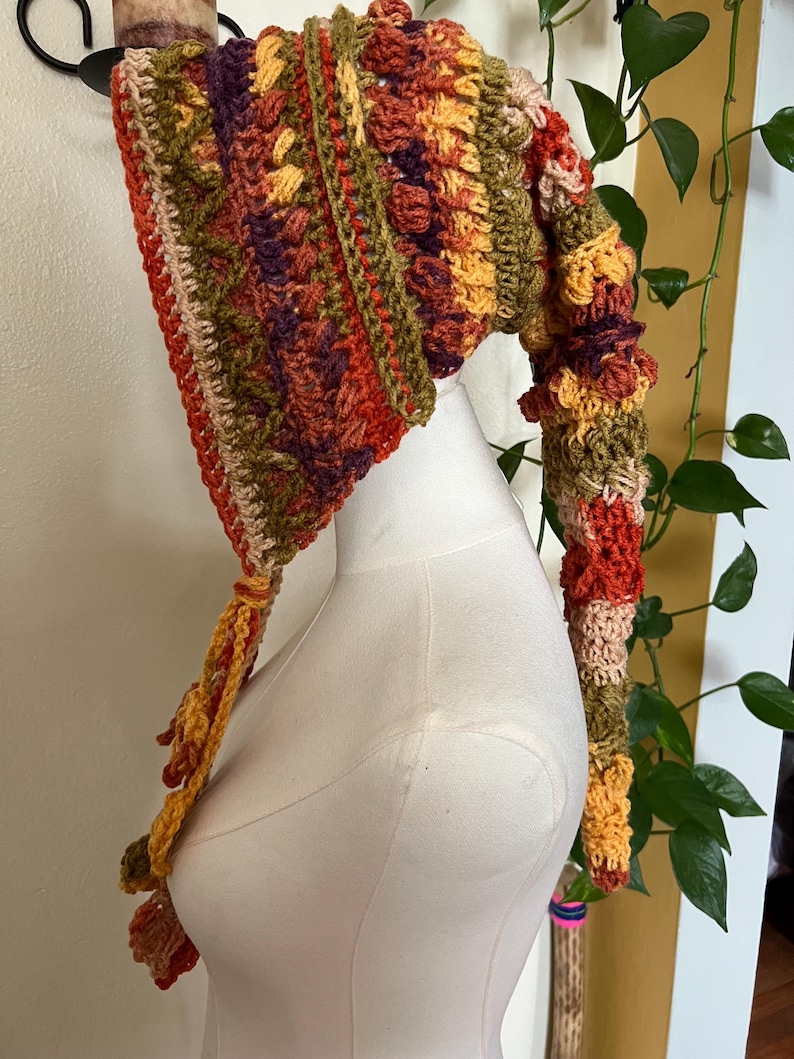 May include: A long, pointed crochet hood featuring a textured, multi-colored pattern in shades of orange, olive green, deep purple, and cream. The headpiece has a tapered, elongated tail and includes braided ties hanging from the front edges.