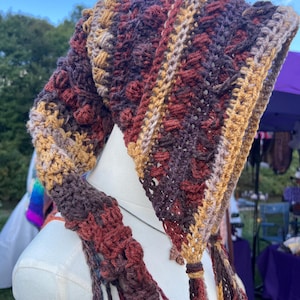 May include: A crochet hood featuring a textured pattern with alternating stripes of deep burgundy, dark brown, and golden yellow yarn. The hood has a pointed shape with long, dangling crocheted ties at the front edges, showcasing intricate stitch work.
