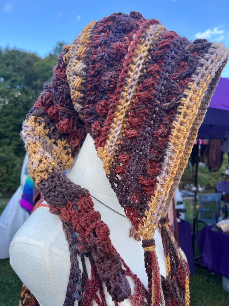 May include: A crochet hood featuring a textured pattern with alternating stripes of deep burgundy, dark brown, and golden yellow yarn. The hood has a pointed shape with long, dangling crocheted ties at the front edges, showcasing intricate stitch work.