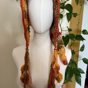 May include: A crochet hood featuring a variegated autumn color palette of orange, yellow, brown, and green yarn. The hood includes long side ties that hang down, each decorated with multiple small, crocheted leaf-shaped embellishments at the ends.