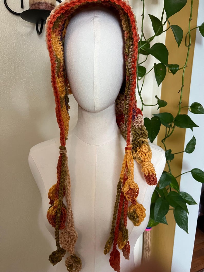 May include: A crochet hood featuring a variegated autumn color palette of orange, yellow, brown, and green yarn. The hood includes long side ties that hang down, each decorated with multiple small, crocheted leaf-shaped embellishments at the ends.