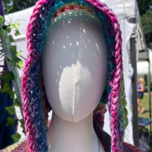 May include: A hooded garment featuring a vibrant, multi-colored crochet pattern with prominent shades of magenta, blue, purple, and green. The hood is constructed with a thick, textured yarn stitch and is displayed on a smooth, white mannequin head.