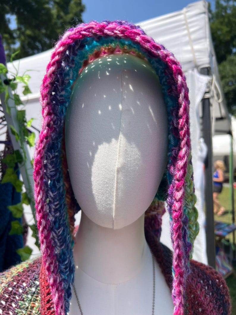 May include: A hooded garment featuring a vibrant, multi-colored crochet pattern with prominent shades of magenta, blue, purple, and green. The hood is constructed with a thick, textured yarn stitch and is displayed on a smooth, white mannequin head.