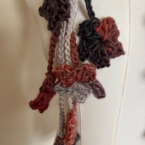 May include: A handmade crochet accessory featuring multiple dangling yarn flowers in shades of dark brown, rust orange, light gray, and charcoal. The floral motifs are connected by thin, braided yarn strands, creating a layered, textured appearance.