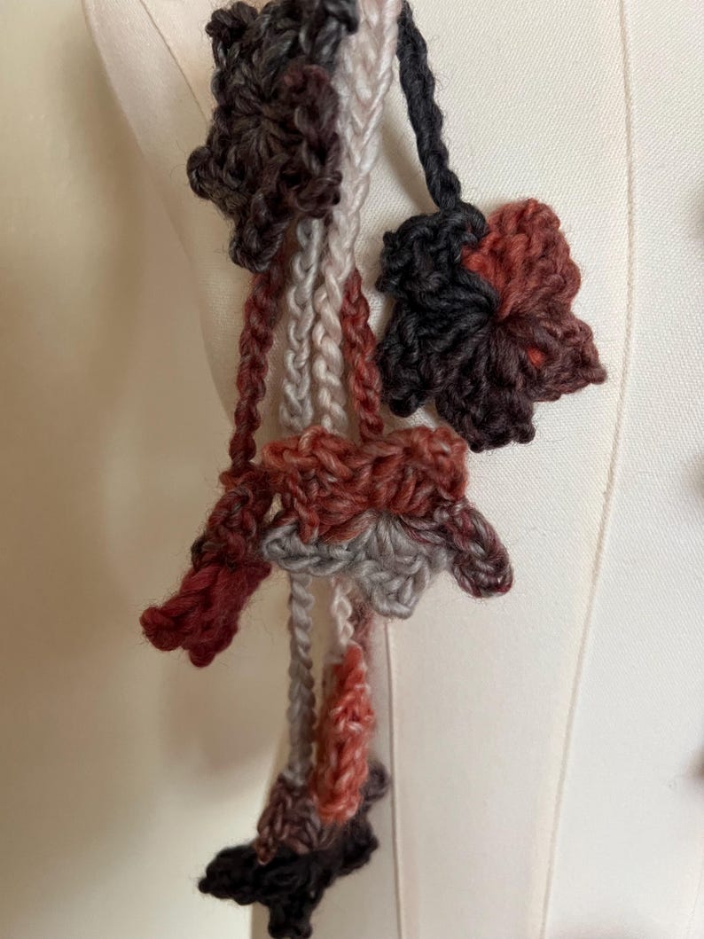 May include: A handmade crochet accessory featuring multiple dangling yarn flowers in shades of dark brown, rust orange, light gray, and charcoal. The floral motifs are connected by thin, braided yarn strands, creating a layered, textured appearance.