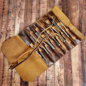 May include: A brown leather roll-up case with a strap for carrying. The case is open and contains a set of 10 wood carving chisels with wooden handles.