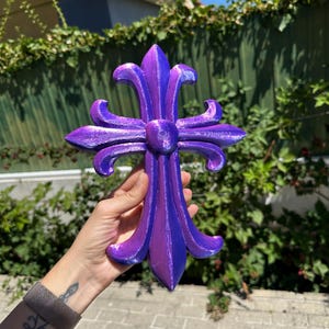 May include: A purple and blue fleur-de-lis cross. The decorative cross is held in front of a green fence and foliage. The cross has a textured surface and a metallic sheen. The design is symmetrical with four stylized petals.