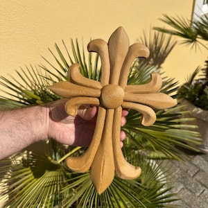 May include: A wooden fleur-de-lis cross in a light brown color. The cross has a detailed design with curved edges and a central circular element. The background features green palm leaves and a yellow wall.