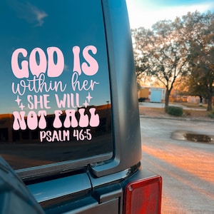 May include: A pink decal on a vehicle window displays the text "GOD IS within her SHE WILL NOT FALL PSALM 46:5". The decal is in a retro font style with star accents. The vehicle is black with a red taillight.