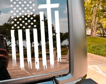 May include: White decal featuring a stylized American flag design with a cross. The flag design includes stars and stripes, with the stripes appearing to drip. The decal is affixed to the rear window of a vehicle.