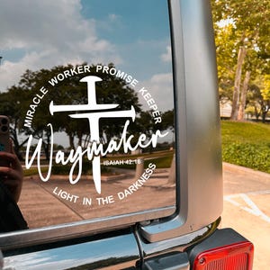 May include: White vinyl decal on a vehicle window. The decal features the word "Waymaker" in a script font, with a cross design and the phrases "Miracle Worker Promise Keeper" and "Light in the Darkness" in a circular arrangement.