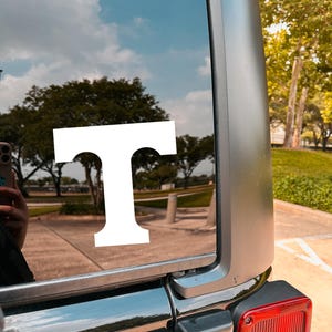 May include: A white vinyl decal in the shape of the letter "T" is affixed to the rear window of a vehicle. The window reflects a blue sky with clouds and a tree-lined landscape. The vehicle's body is dark gray.