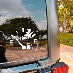 May include: White vinyl decal of a fuel gauge with a witch on a broomstick, the needle pointing to empty. The gauge is on a car window, with a reflection of a blue sky and trees.