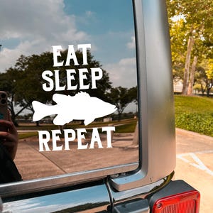 May include: White vinyl decal applied to a vehicle window. The decal features the words "EAT SLEEP REPEAT" in a bold, sans-serif font, with a white fish silhouette between "SLEEP" and "REPEAT". The background shows a cloudy sky and trees.
