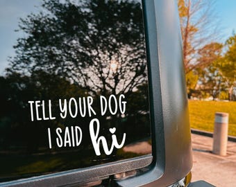 May include: A white vinyl sticker on a car window displays the text "TELL YOUR DOG I SAID hi" in a whimsical font. The sticker is affixed to a black vehicle, with a blurred backdrop of trees and a blue sky.