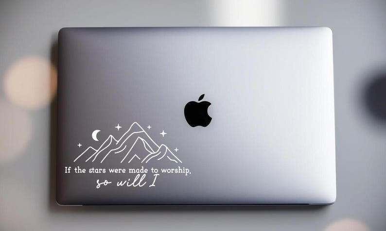 May include: A silver laptop with a white sticker featuring a mountain range, a crescent moon, and stars. The sticker also includes the text "If the stars were made to worship, so will I."