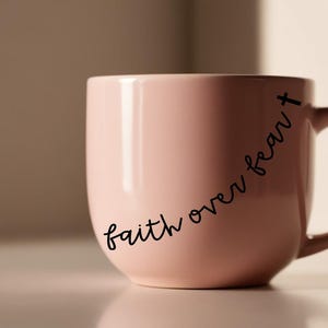 May include: A light pink ceramic mug with a curved handle. The mug features the phrase "faith over fear" in a black, cursive font. A small cross is at the end of the phrase.