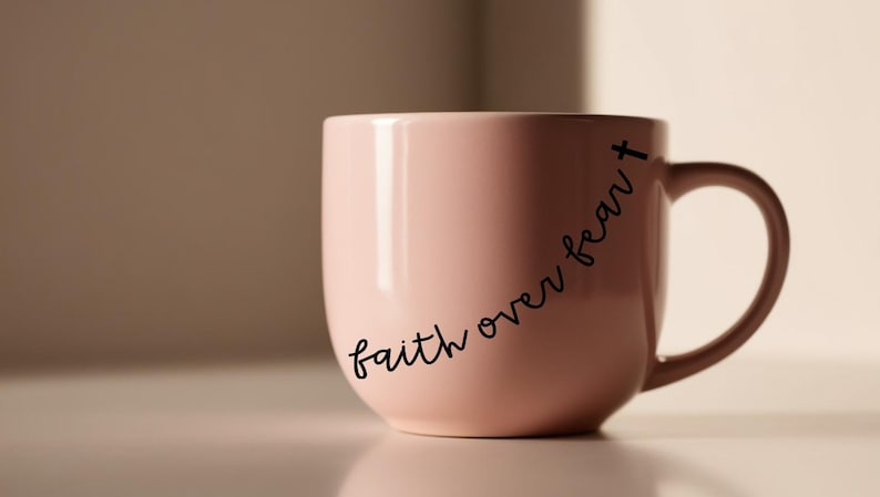 May include: A light pink ceramic mug with a curved handle. The mug features the phrase "faith over fear" in a black, cursive font. A small cross is at the end of the phrase.