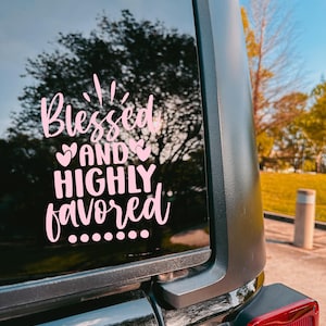 May include: Pink vinyl decal applied to a vehicle window. The decal features the words "Blessed and Highly Favored" in a cursive font, with decorative hearts and dots. The background shows a tree and a blue sky.